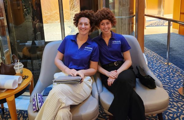 Leah Meek and Katie Dinkel sitting on a cruise deck waiting to present in Bob Ross Wigs