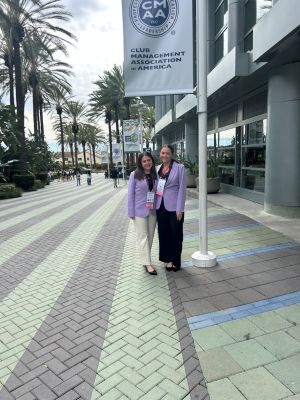 Sophia Struck and Jojo Valdivia posing on a brick street under CMAA World Conference Banner with palm trees and buildings in the background